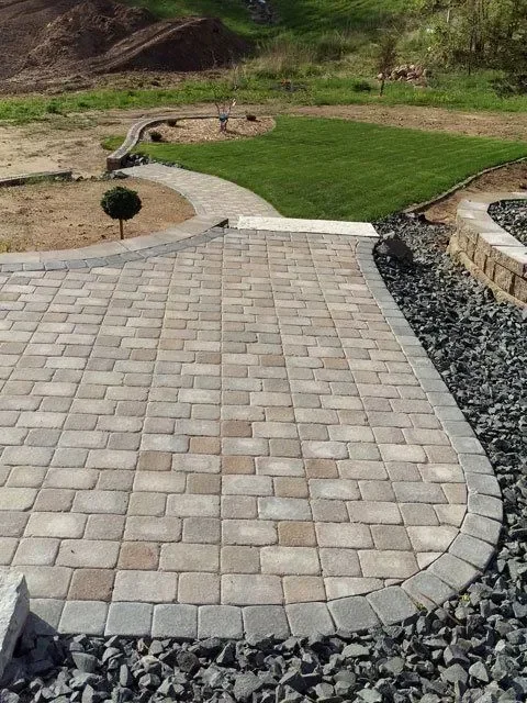 Curved tan paver patio with black rock borders and adjacent green lawn installation