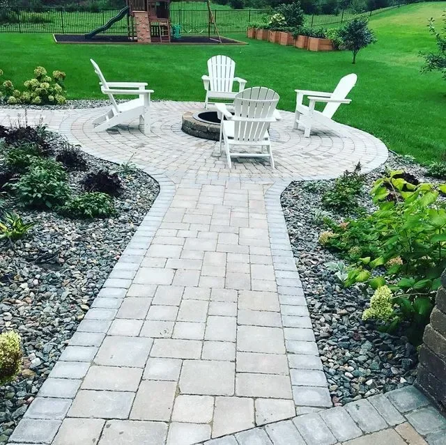 Circular paver patio with fire pit, white Adirondack chairs, gravel beds, and connecting walkway