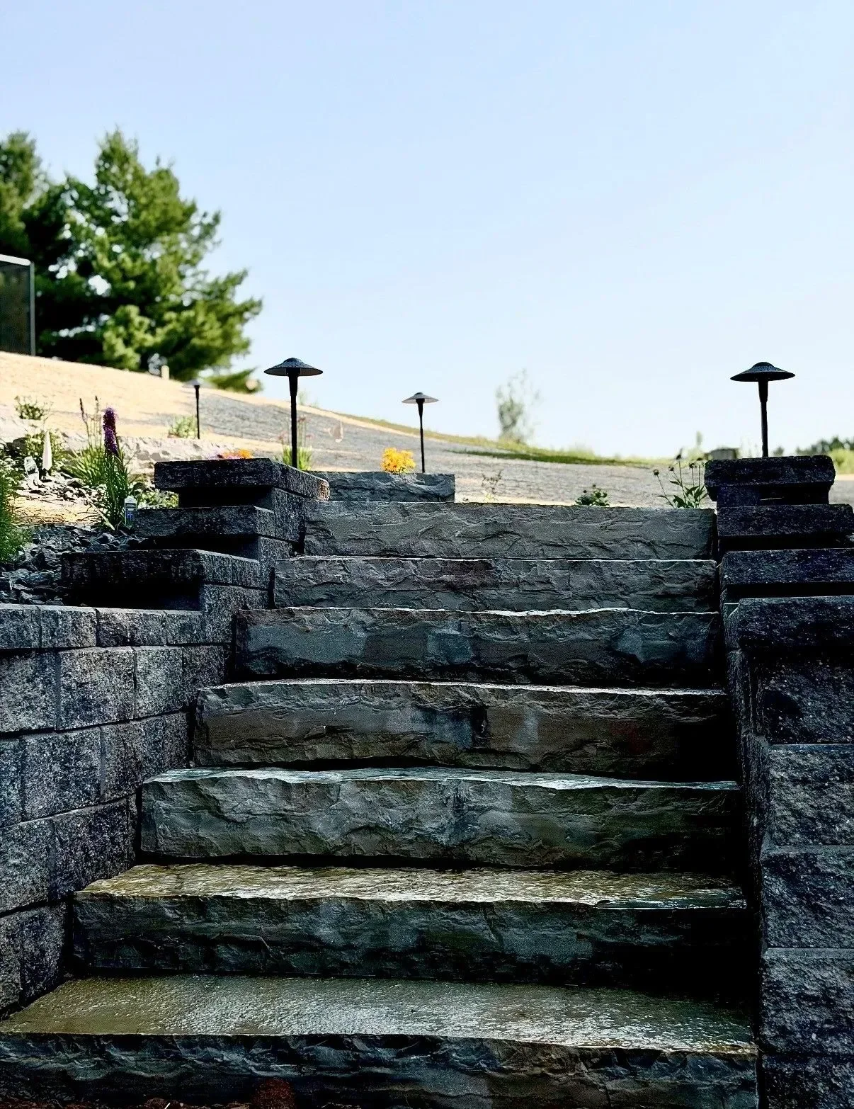 Tiered natural stone retaining wall with steps, landscape lighting, and planted beds on sloped terrain