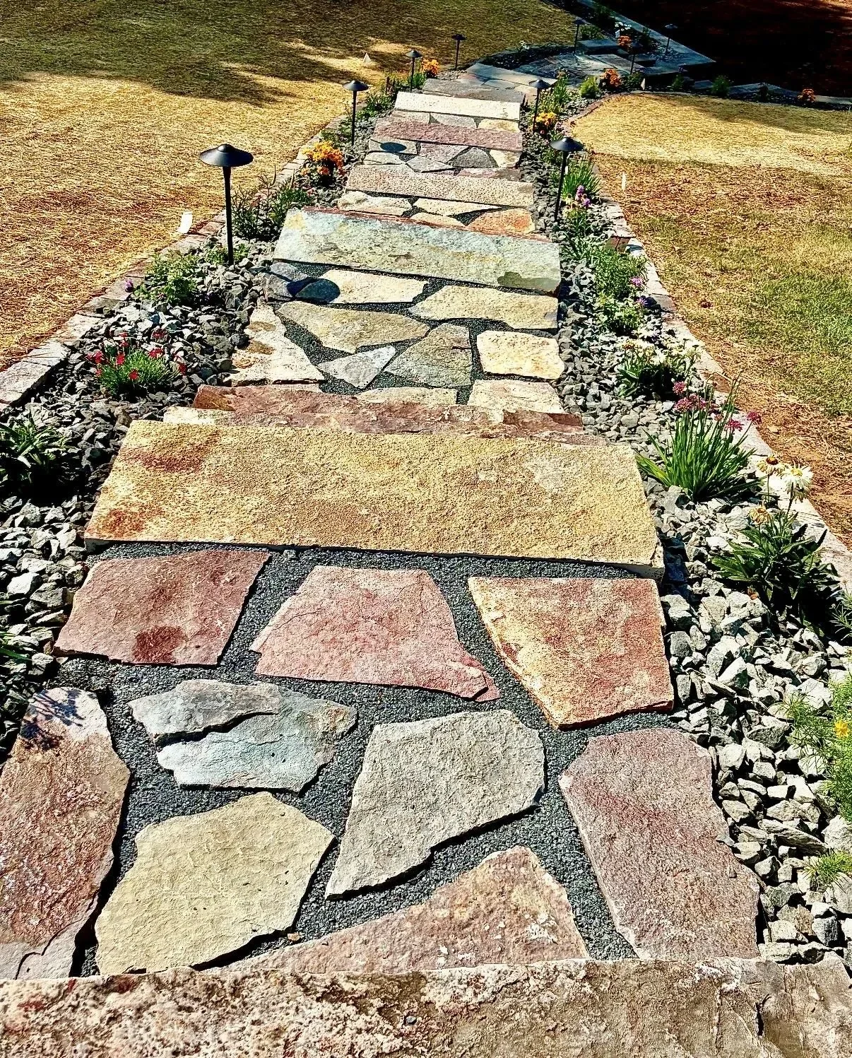 Flagstone pathway with mixed-color stone pavers, dark aggregate base, and planted borders with river rock