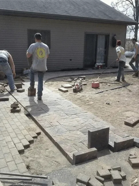 Workers installing interlocking pavers on residential patio with concrete edging and steps