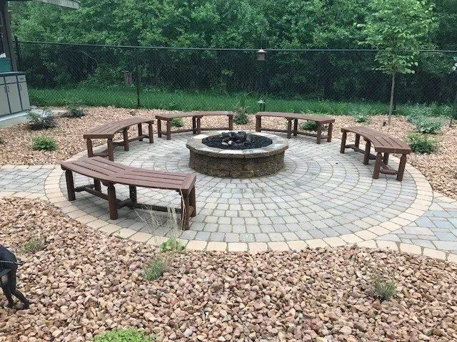 Circular paver patio with central fire pit, wooden benches, and river rock landscaping