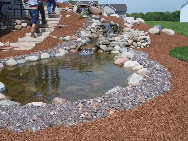 Backyard pond with stone border, river rock edging, and mulch landscaping on sloped terrain